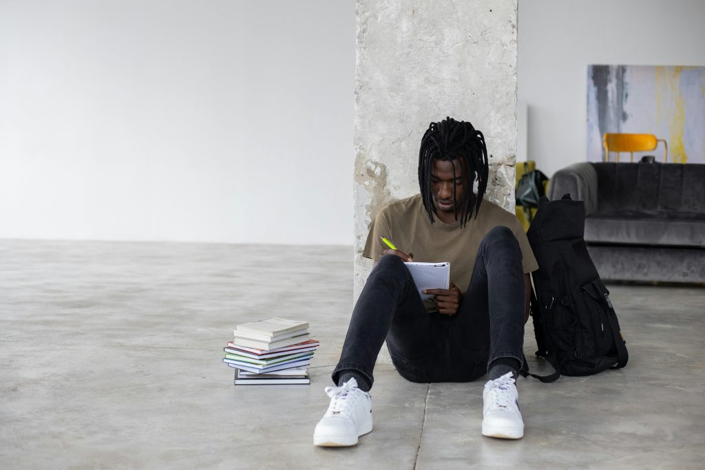 Young man sitting on the floor, studying with notebooks and backpack indoors.