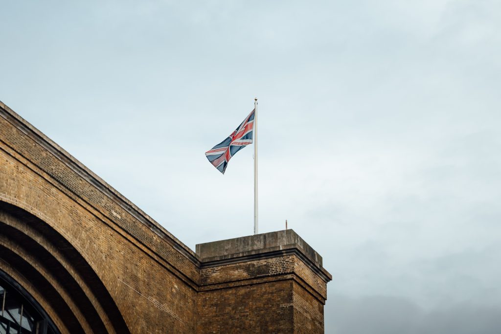 Union Jack flag waving atop a historic brick building in London, UK, against a cloudy sky.